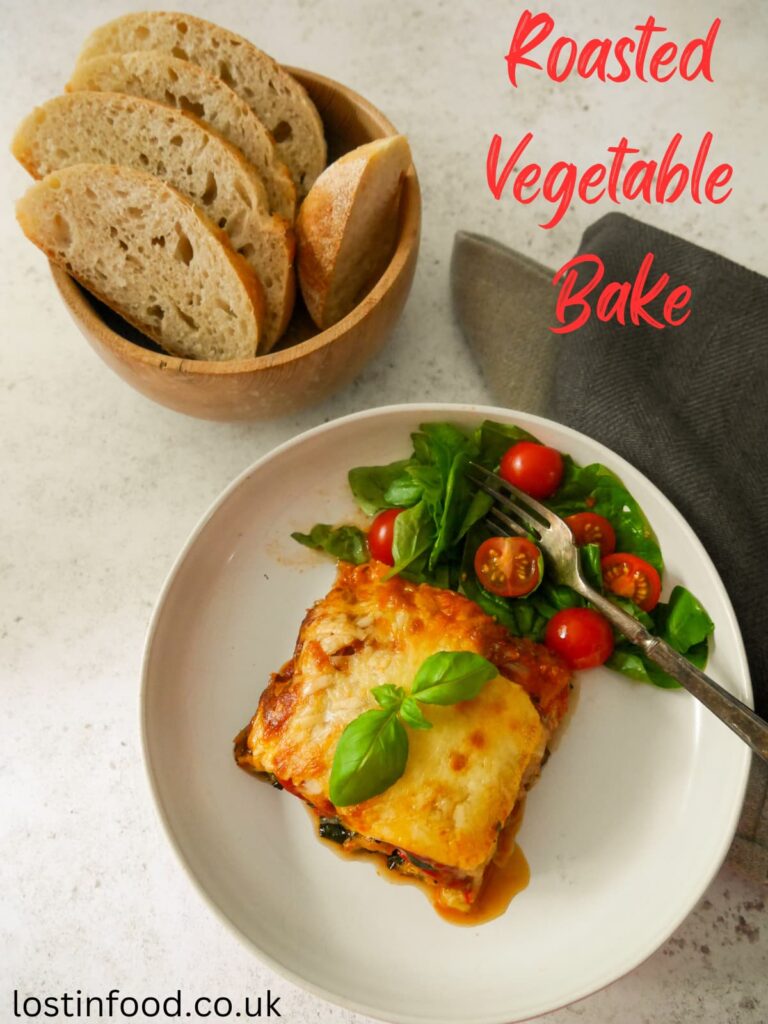 A pinnable image with recipe title and white plate with a wedge of roasted vegetable bake, garnished with fresh basil, with a wooden bowl of sliced sourdough bread set alongside.