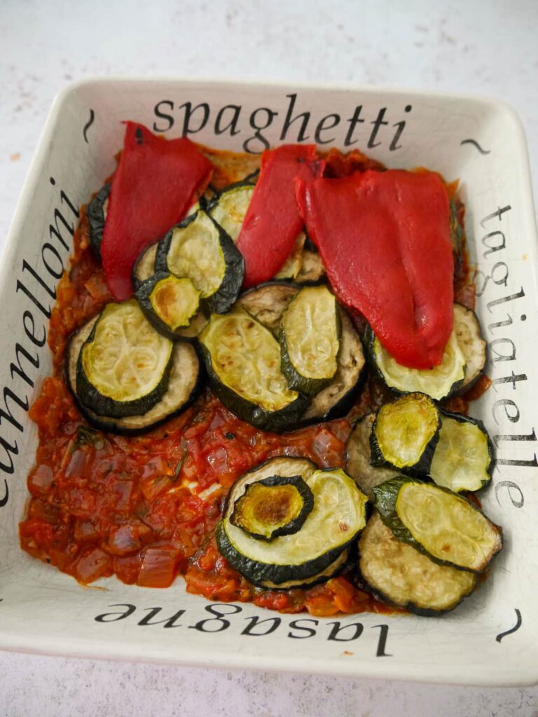 A baking dish spread with tomato and basil sauce, topped with slices of roasted aubergine, courgette and red bell pepper.