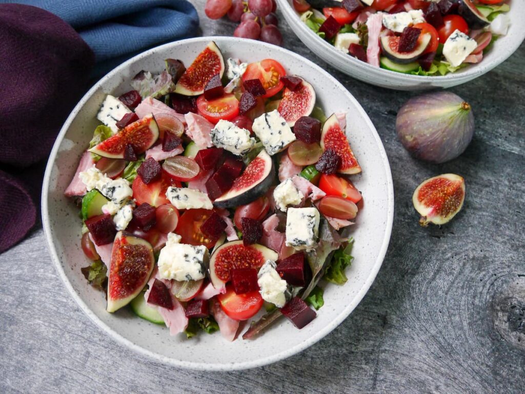 Two white bowls filled with ham salad with figs and blue cheese with a small bunch of red grapes, a whole fig and sliced fig set alongside.