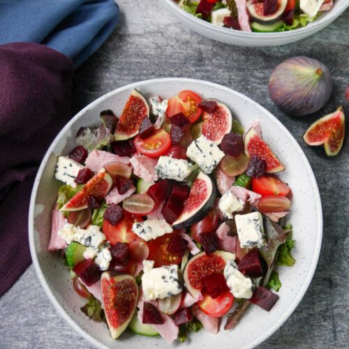 Two white bowls filled with ham salad with figs and blue cheese with a whole fig and sliced fig set alongside.