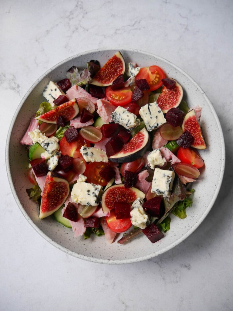 A bowl filled with ham, blue cheese, figs, red grapes, beetroot, cucumber and lettuce.