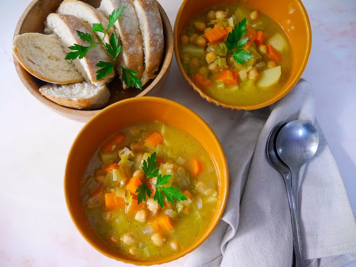 Two bowls of leek, potato and chickpea soup with a bowl of sliced bread set alongside.