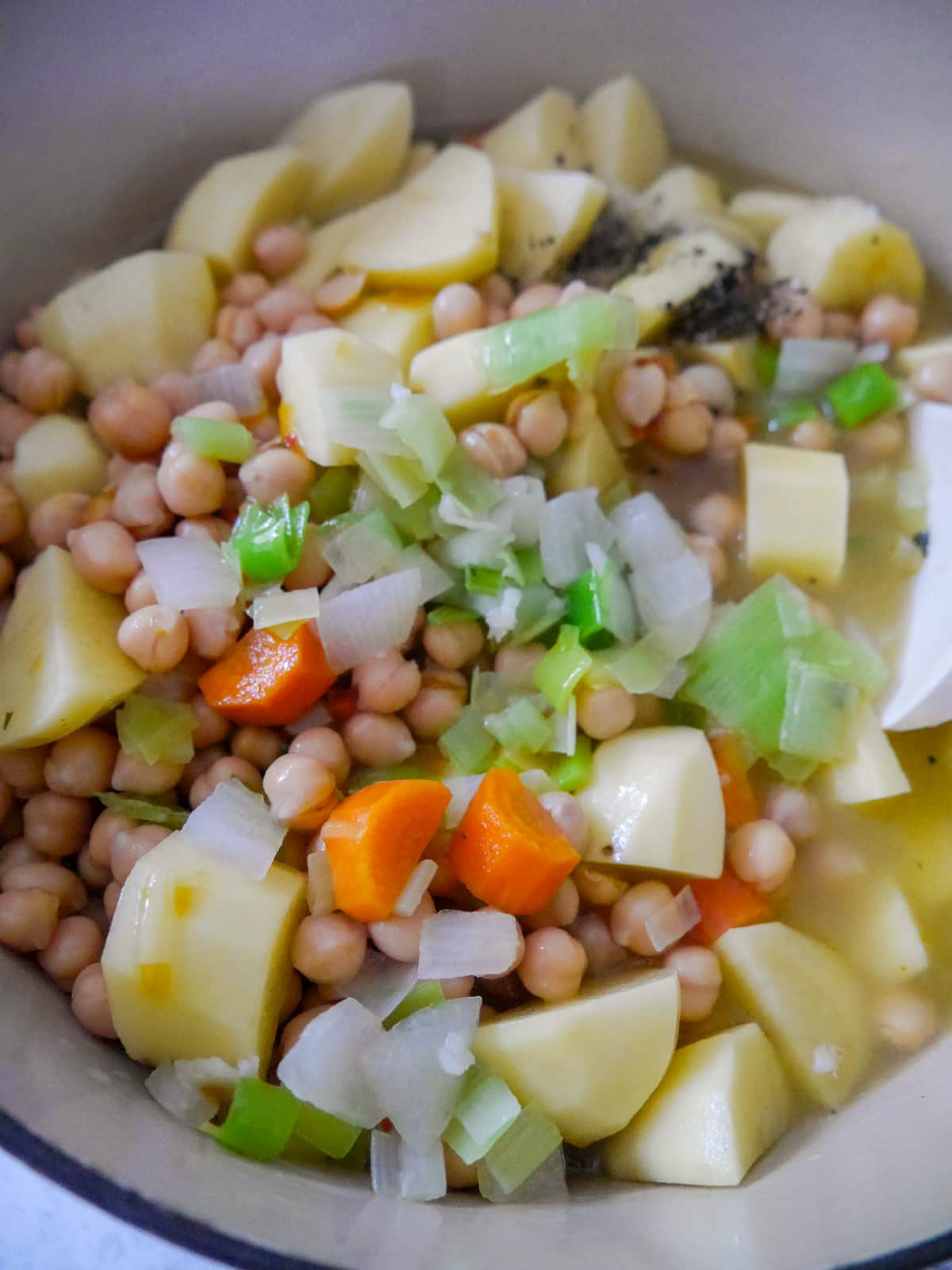 A large pan filled with sauteed vegetables, with chickpeas, potatoes and added black pepper and vegetable stock.