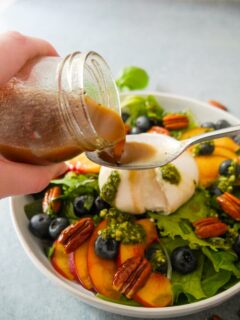 Lemon balsamic dressing being poured over a salad of sliced peaches, burrata cheese, blueberries, candied pecans and pesto.