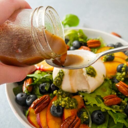 Lemon balsamic dressing being poured over a salad of sliced peaches, burrata cheese, blueberries, candied pecans and pesto.