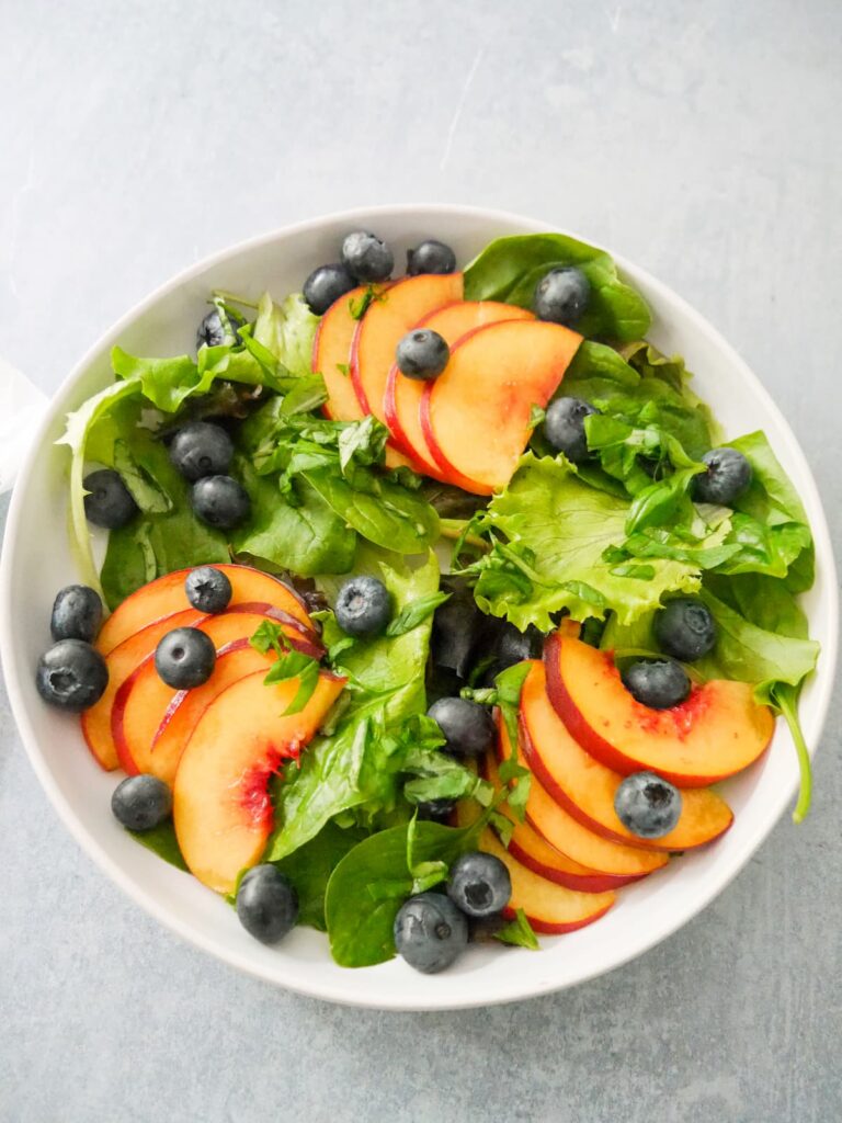 A bowl filled with mixed green salad leaves, sliced peaches and blueberries.