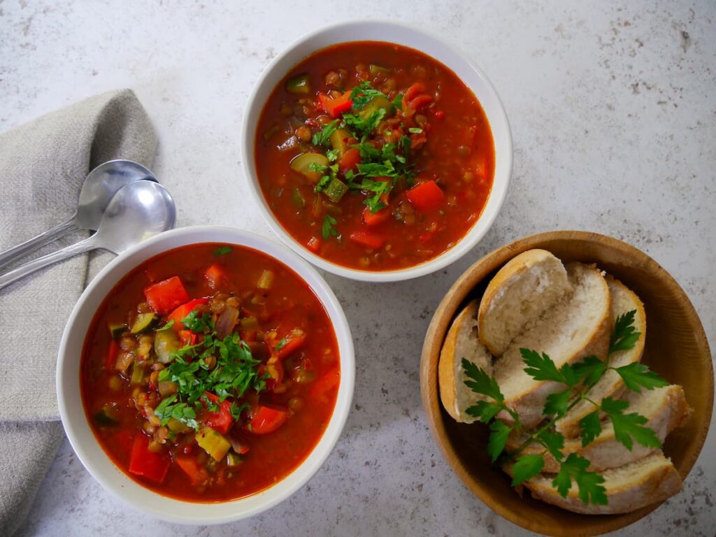 Two bowls of tomato and vegetable soup garnished with fresh parsley, with a bowl of sliced crusty bread set alongside.