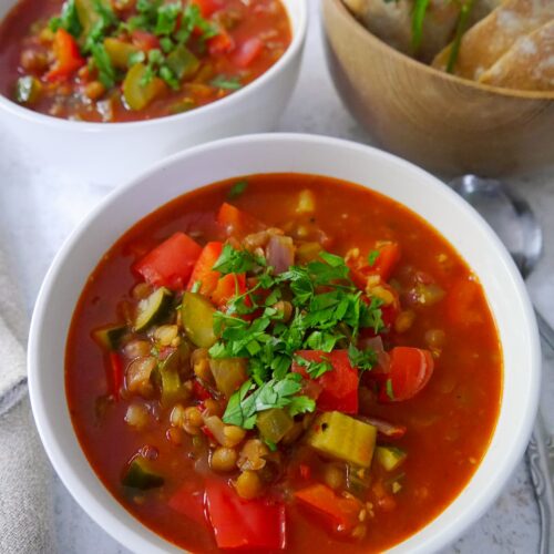 Two bowls of tomato and vegetable soup garnished with fresh parsley, with a bowl of sliced crusty bread set alongside.