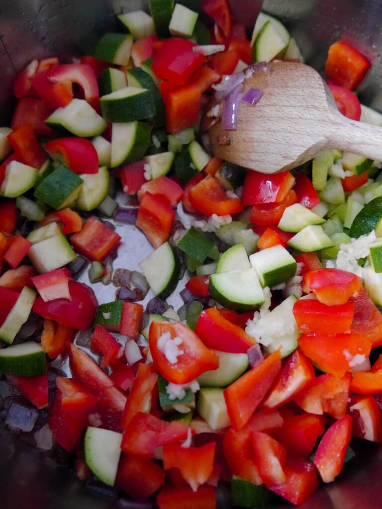A large saucepan filled with sauteed red onions with added red pepper, courgettes and garlic.