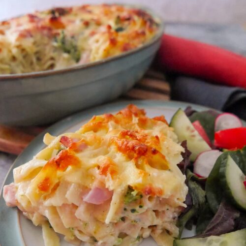 A plate with a serving of ham and broccoli pasta bake with salad, and a dish of pasta bake set alongside.