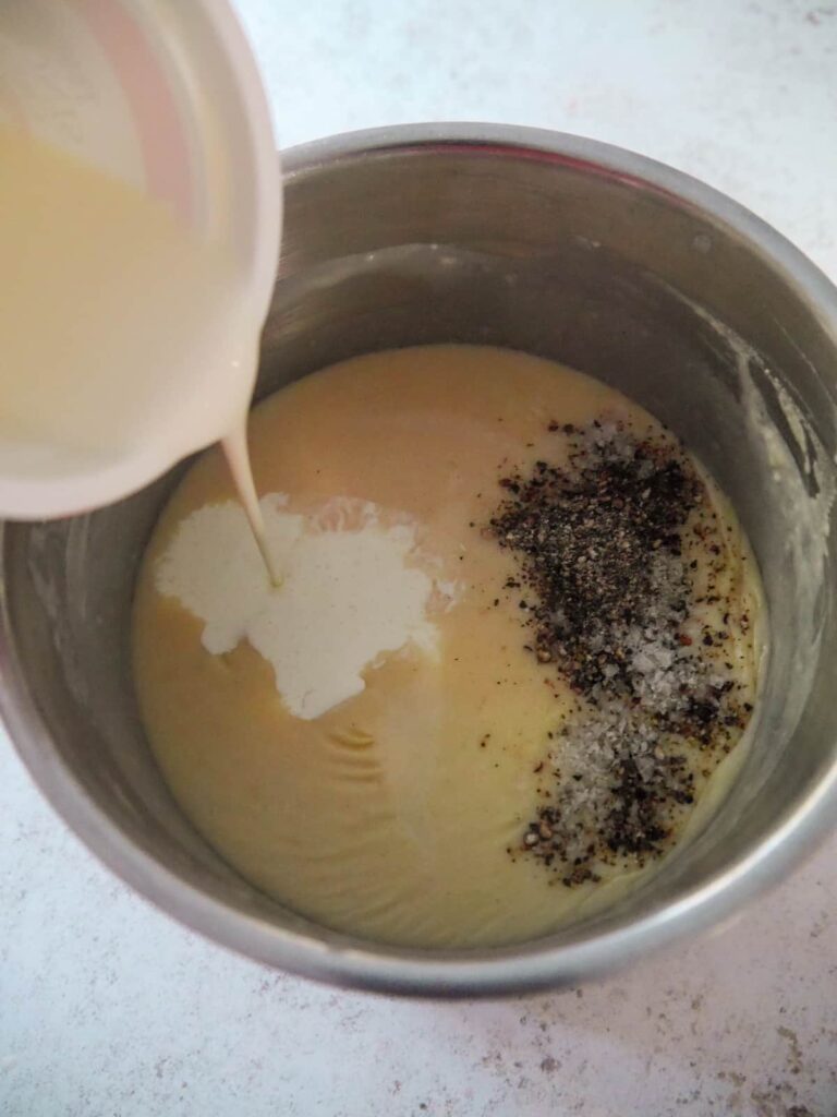 A white sauce base with salt and pepper and double cream being poured added to saucepan.