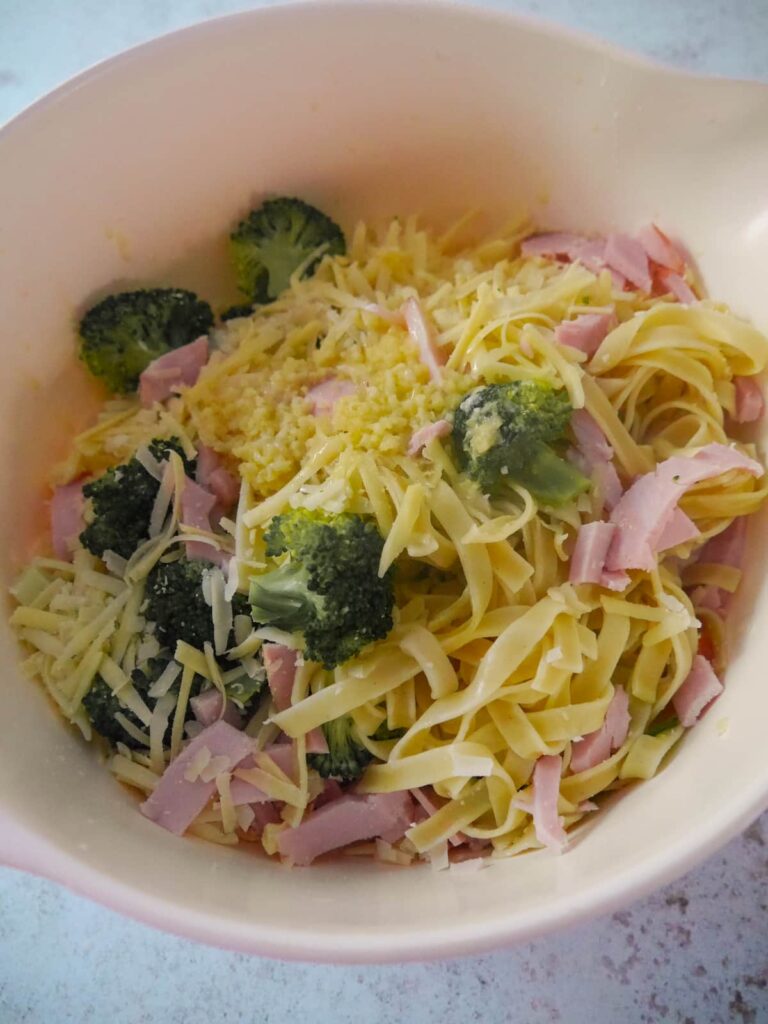 A large bowl filled with cooked tagliatelle pasta, strips of cooked ham, par-cooked broccoli florets and grated cheese.
