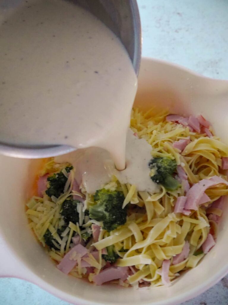 Creamy white sauce being poured over a large bowl filled with cooked tagliatelle pasta, strips of cooked ham, par-cooked broccoli florets and grated cheese.