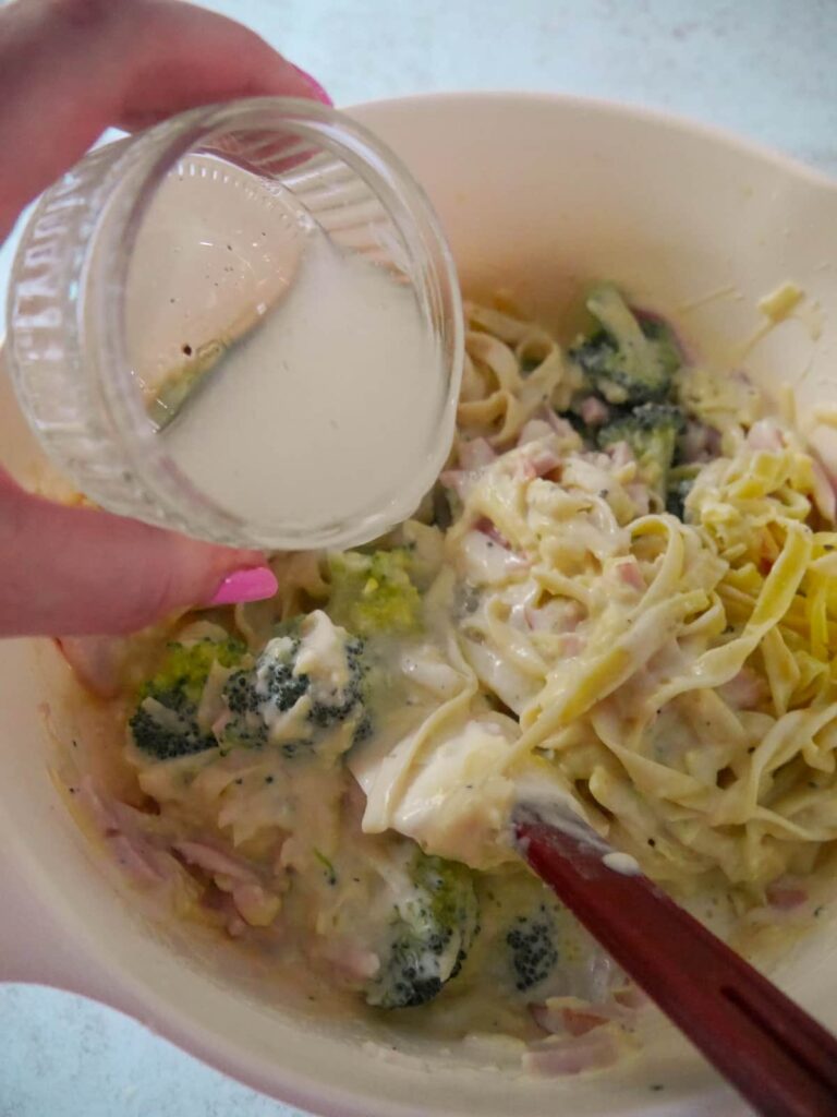 A cup of reserved pasta cooking water being added to the bowl of ham and broccoli pasta bake ingredients.