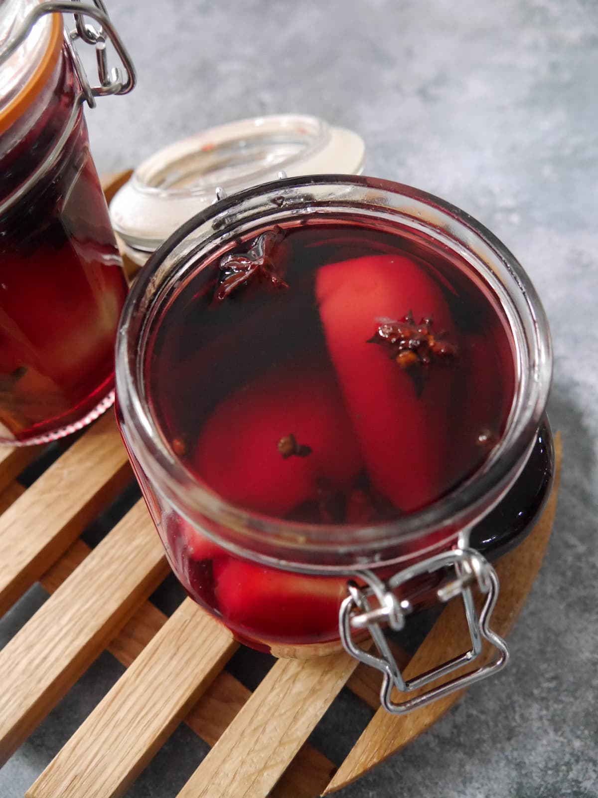Two glass jars filled with pears covered in red wine with whole spices, set on a wooden trivet.