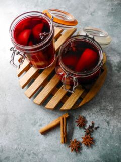 Two glass jars filled with pears covered in red wine with whole spices, set on a wooden trivet with whole cloves, star anise and cinnamon sticks set alongside.