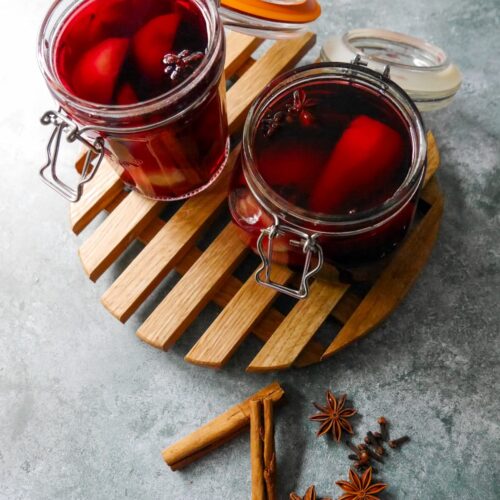Two glass jars filled with pears covered in red wine with whole spices, set on a wooden trivet with whole cloves, star anise and cinnamon sticks set alongside.