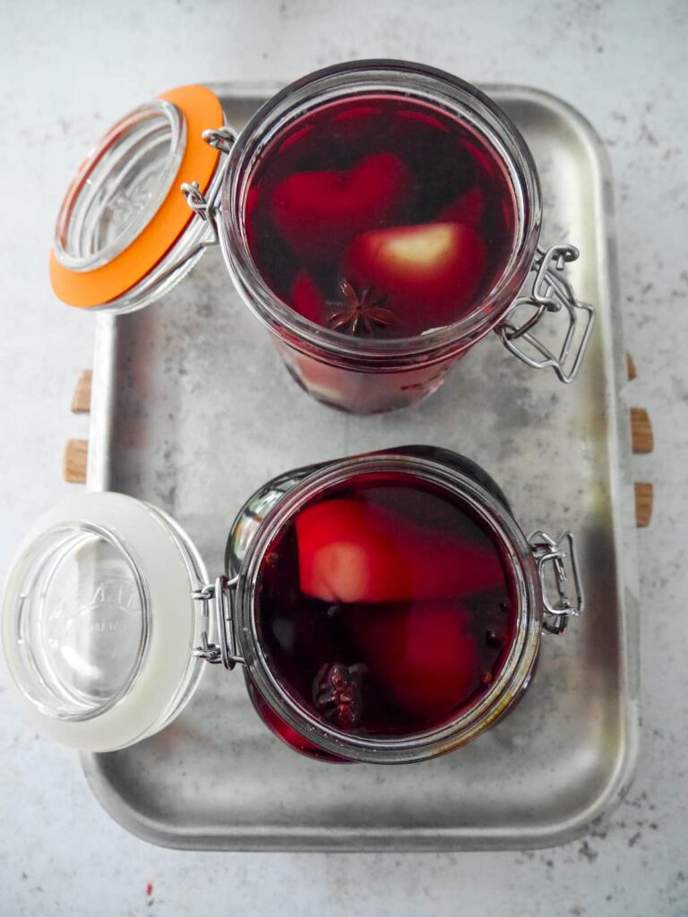 Two glass jars filled with pears covered in red wine with whole cloves, star anise and cinnamon sticks.