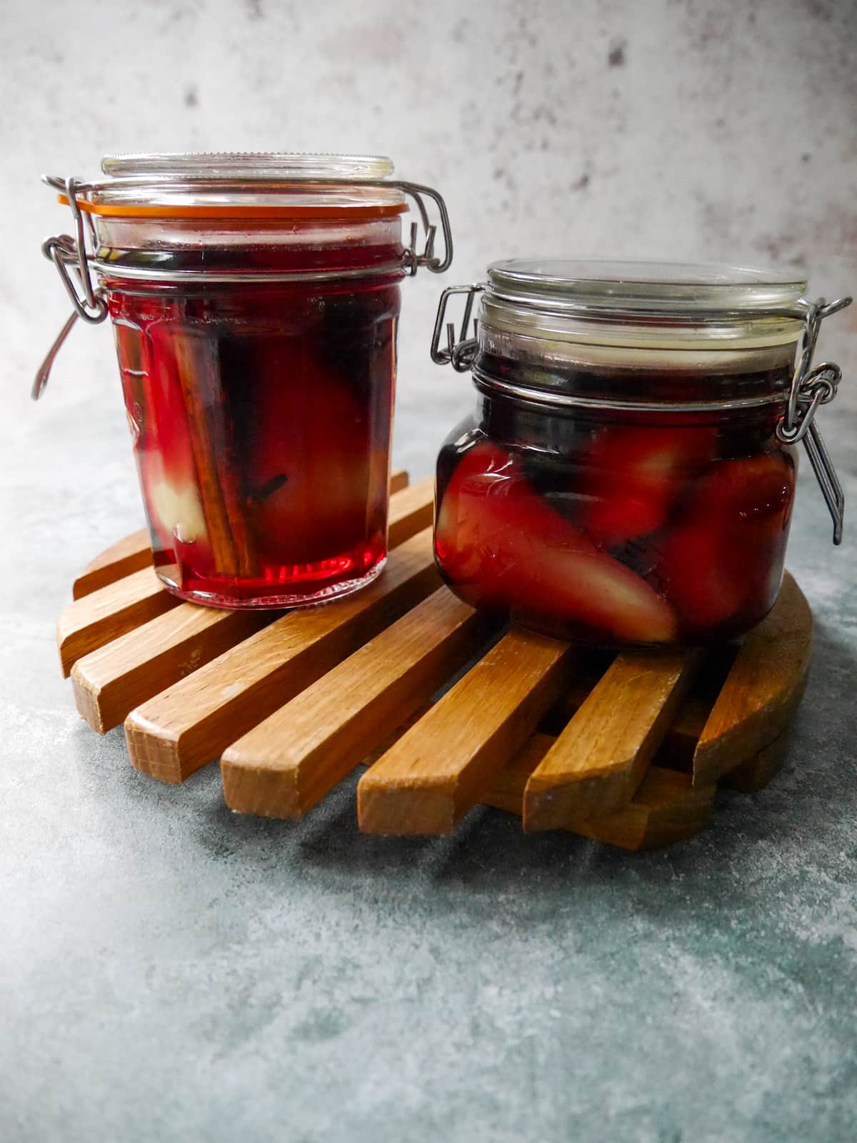 Two glass jars filled with pears covered in red wine with whole spices, set on a wooden trivet.