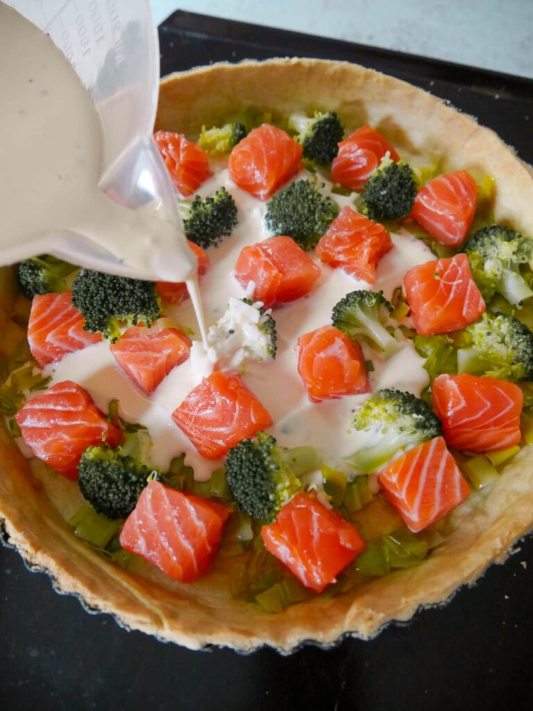 Part baked pastry tin with a layer of sauteed leeks, topped with chunks of raw salmon and par-cooked broccoli florets with egg, cream and cheese mixture being poured over.