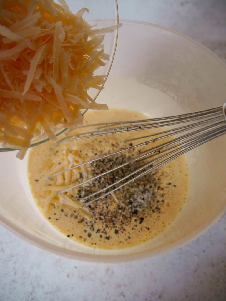 A measuring jug filled with mixed eggs and cream with salt and pepper and grated cheese being added.