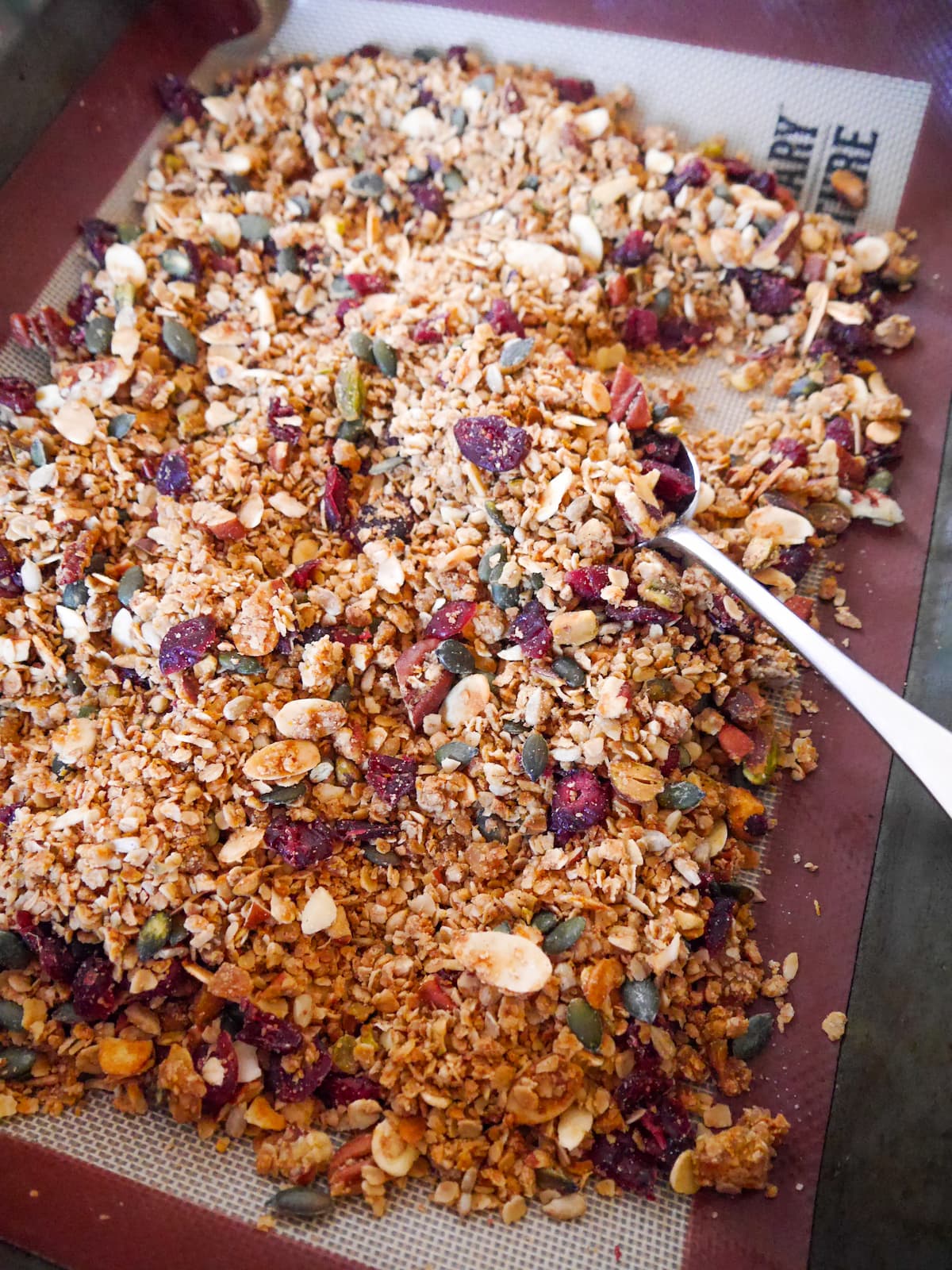 A roasting dish with Christmas spiced granola with orange and cranberries being stirred together.
