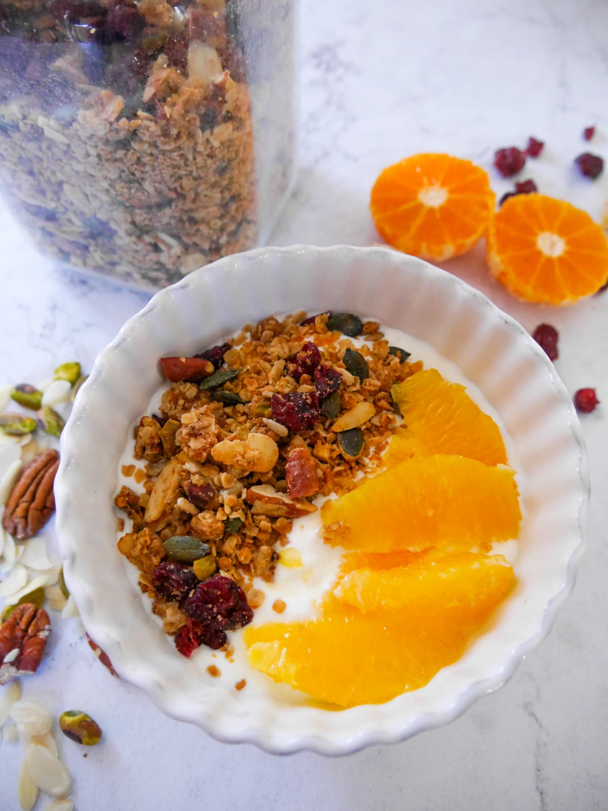 A white bowl filled with Greek yogurt topped with slices of fresh orange and Christmas spiced granola with orange and cranberries, with a jar of granola set alongside.