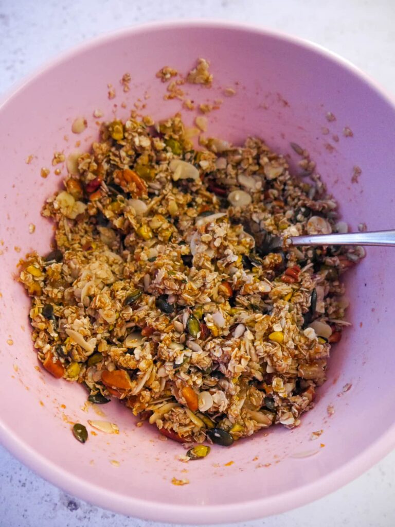 A pink bowl with wet and dry granola ingredients being stirred together.