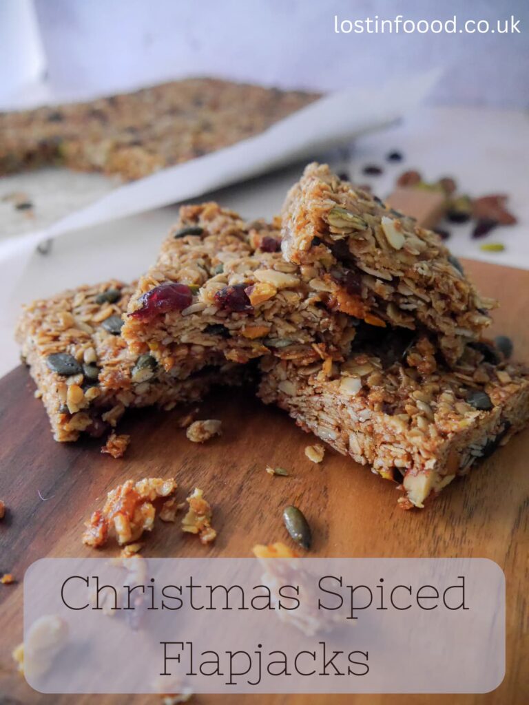Pinnable image with recipe title and wooden board topped with 5 slices of Christmas spiced flapjacks with a wire tray of cut flapjacks set alongside.