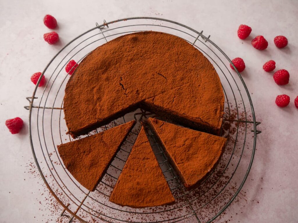 A flourless chocolate cake, sliced and topped with a dusting of cocoa powder set on a wire rack.