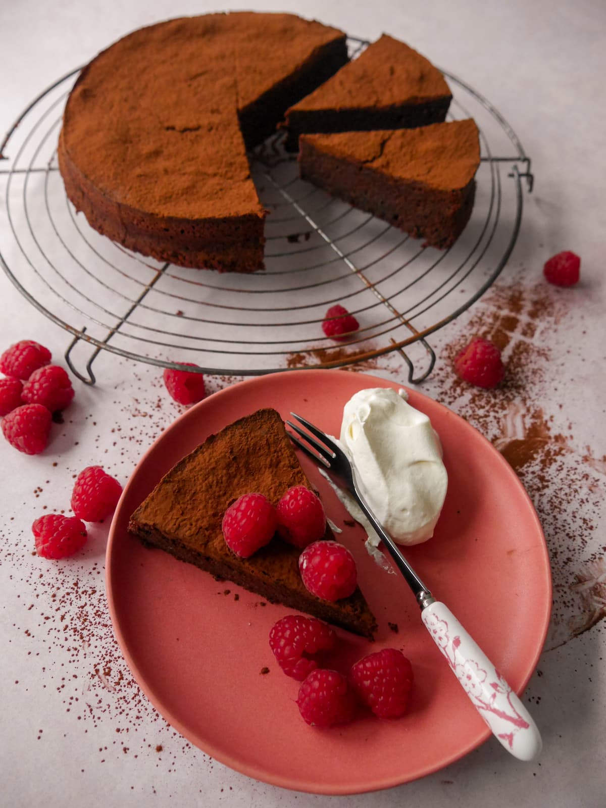 A flourless chocolate cake with amaretto sliced and set on a wire rack, with a pink plate with a slice of flourless chocolate amaretto cake topped with fresh raspberries and served with whipped cream set alongside.