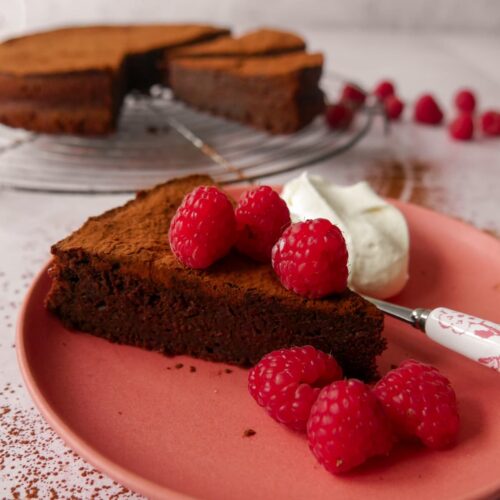 A flourless chocolate cake with amaretto sliced and set on a wire rack, with a pink plate with a slice of flourless chocolate amaretto cake topped with fresh raspberries and served with whipped cream set alongside.