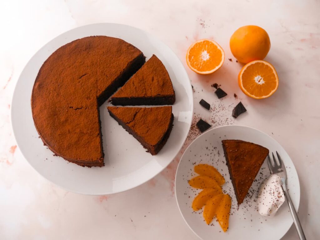 A white plate with a sliced flourless chocolate orange cake with a serving of cake with orange slices and whipped cream set alongside.