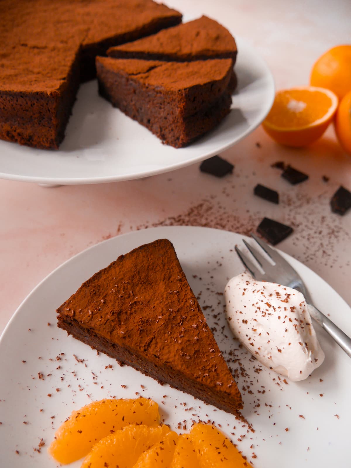 A white plate with a sliced flourless chocolate orange cake with a serving of cake with orange slices and whipped cream set alongside.