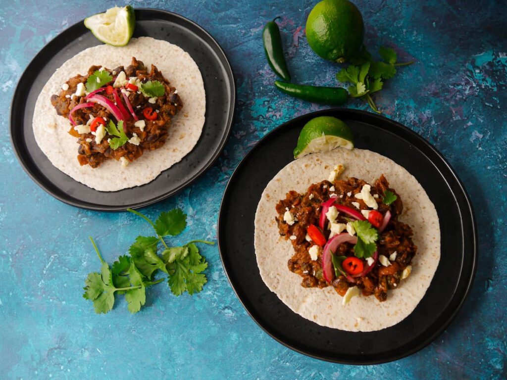 Two black plates with a flour tortilla topped with refried beans garnished with pickled red onions, cheese, sliced red chillies, coriander leaves and a wedge of lime, with coriander, a whole lime and 2 jalapeno chilli peppers set alongside.