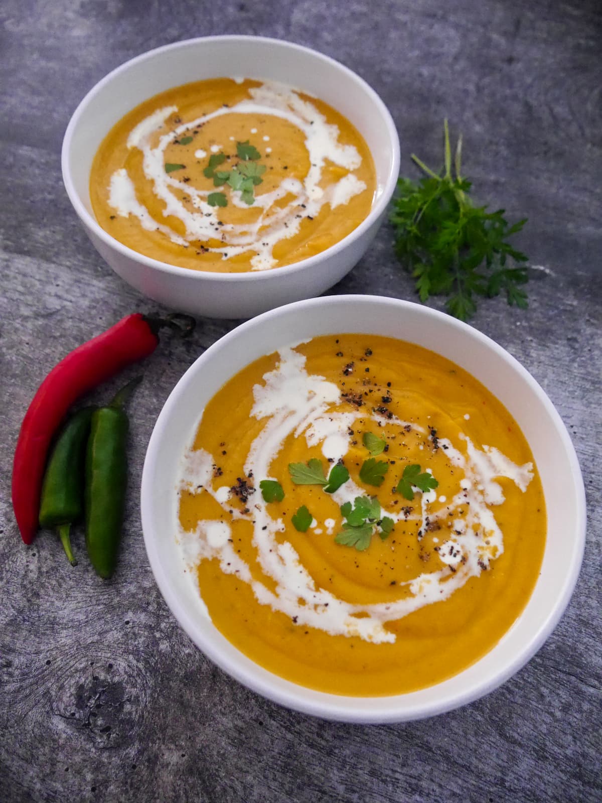 Two white bowls filled with sweet potato, coconut and chilli soup garnished with a drizzle of coconut milk and parsley.