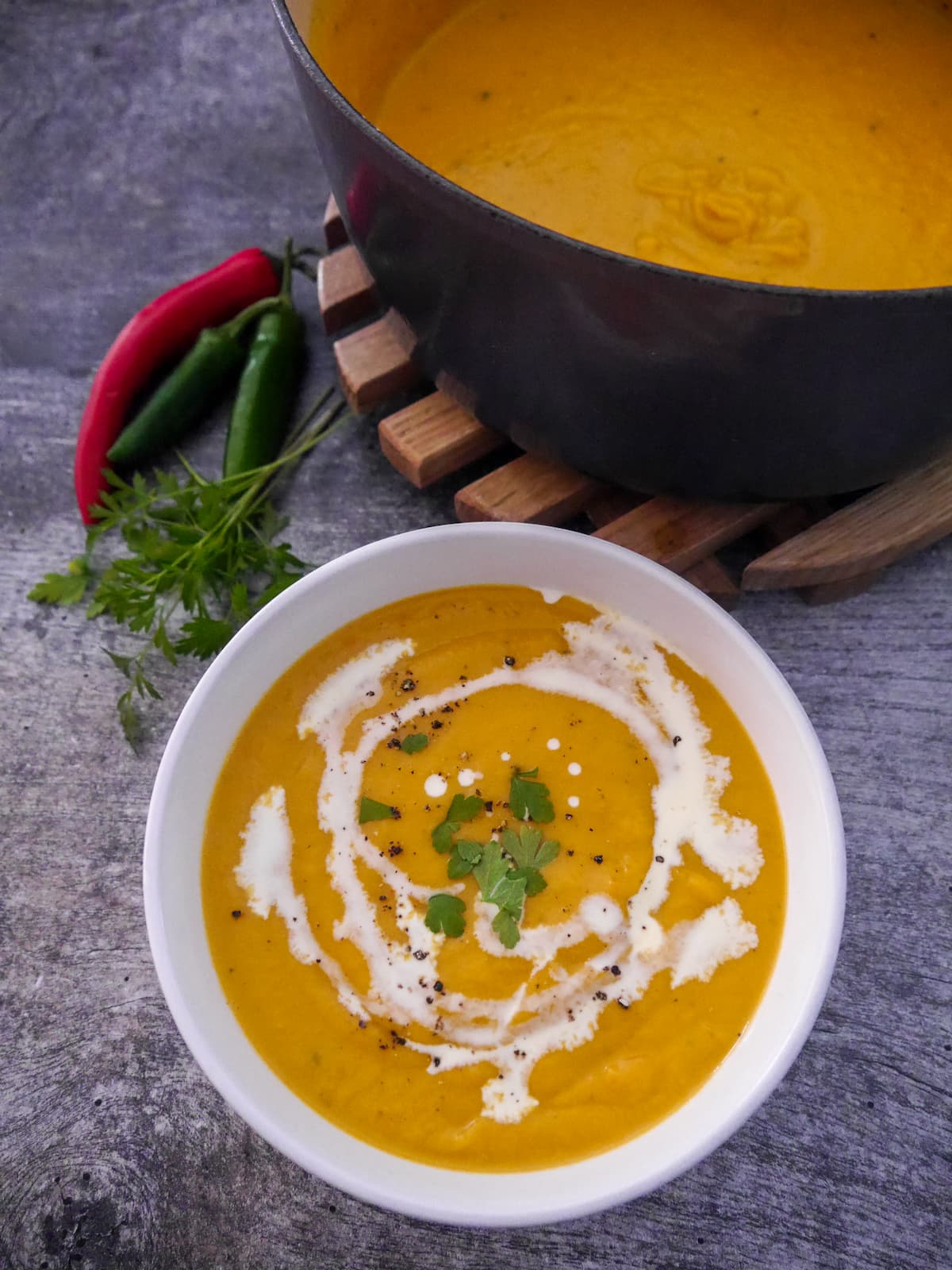 White bowl filled with sweet potato, coconut and chilli soup garnished with a drizzle of coconut milk and parsley, with a Dutch oven filled with soup set alongside.