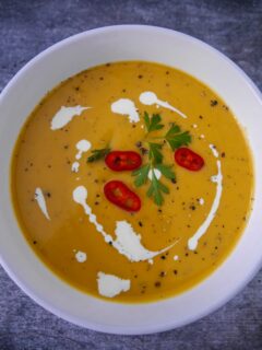 White bowl filled with sweet potato, coconut and chilli soup garnished with a drizzle of coconut milk, sliced red chillies and parsley.