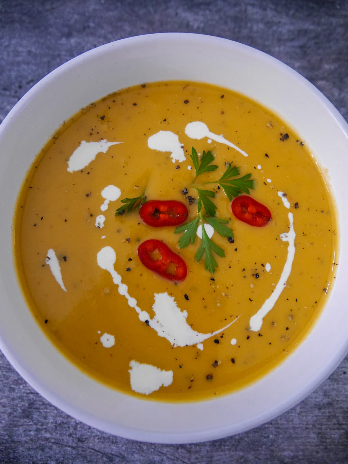 White bowl filled with sweet potato, coconut and chilli soup garnished with a drizzle of coconut milk, sliced red chillies and parsley.