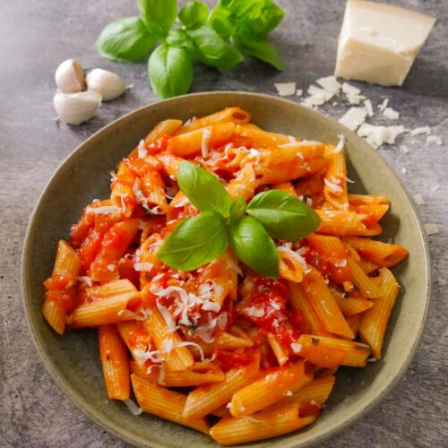 A green bowl filled with penne pasta in a tomato, basil pasta sauce with a garnish of parmesan cheese and fresh basil leaves, with a block of parmesan, fresh basil leaves and garlic set alongside.
