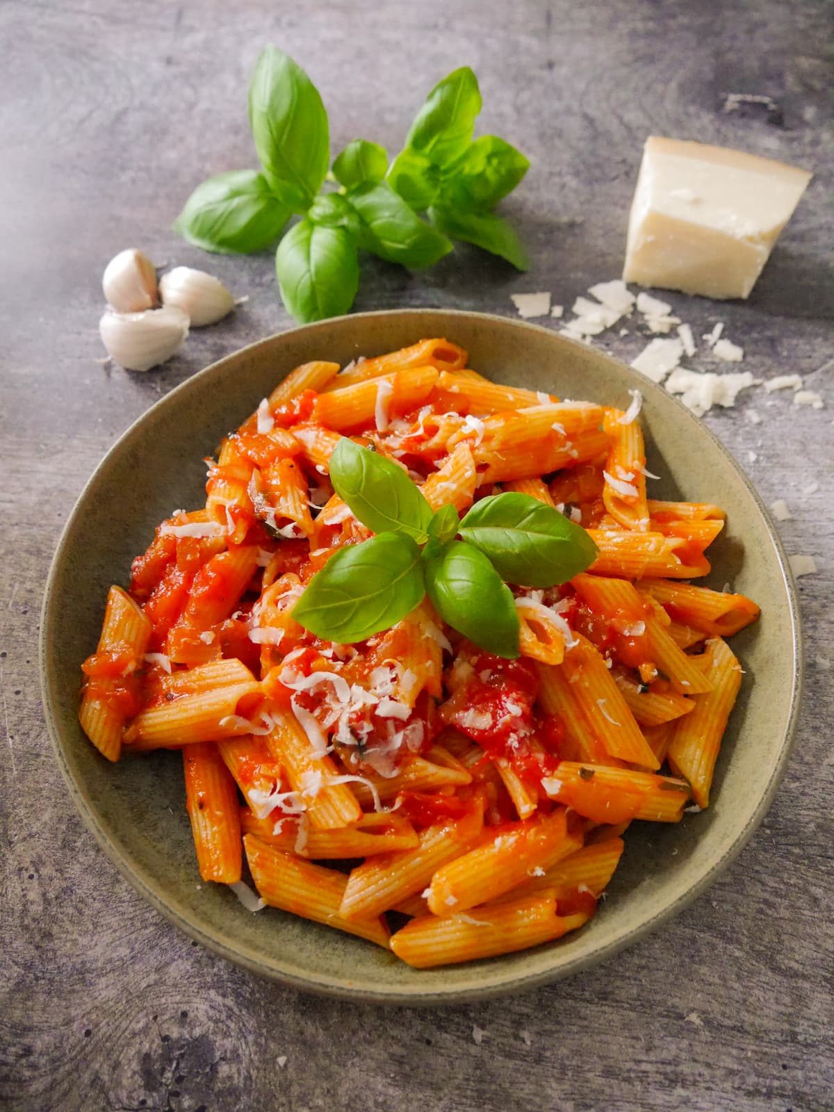 A green bowl filled with penne pasta in a tomato, basil pasta sauce with a garnish of parmesan cheese and fresh basil leaves, with a block of parmesan, fresh basil leaves and garlic set alongside.