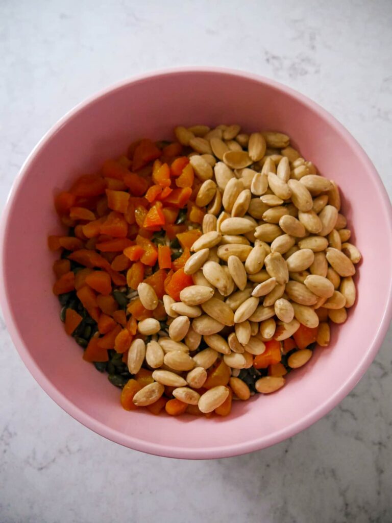 A large pink mixing bowl filled with chopped dried apricots, whole roasted almonds, seeds and rolled oats.
