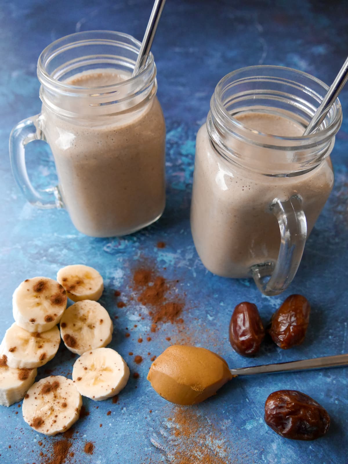 2 glasses filled with chocolate peanut butter smoothie with sliced bananas, whole dates and a spoon of peanut butter set alongside.
