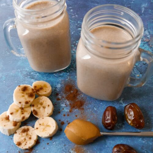 2 glasses filled with chocolate peanut butter smoothie with sliced bananas, whole dates and a spoon of peanut butter set alongside.
