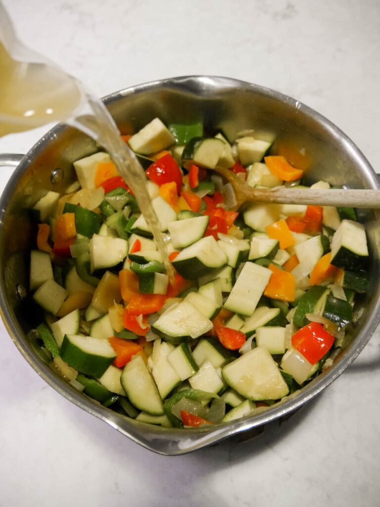A pan filled with sauteed vegetables with vegetable stock being poured in.