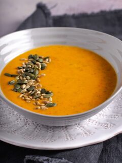 White bowl of courgette and pepper soup, topped with toasted seeds and set on a white plate.
