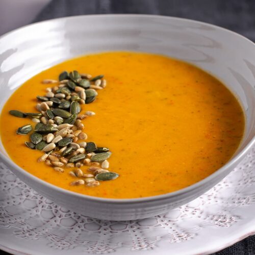White bowl of courgette and pepper soup, topped with toasted seeds and set on a white plate.