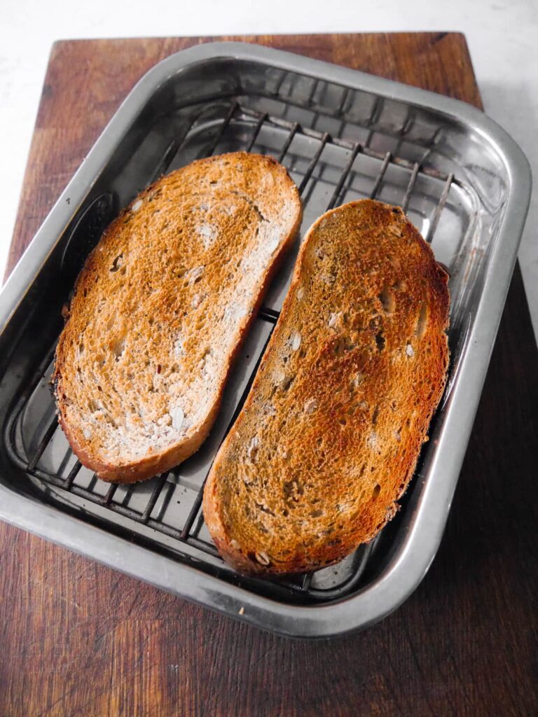 A baking tray with wire rack topped with 2 slices of toasted seeded bread.