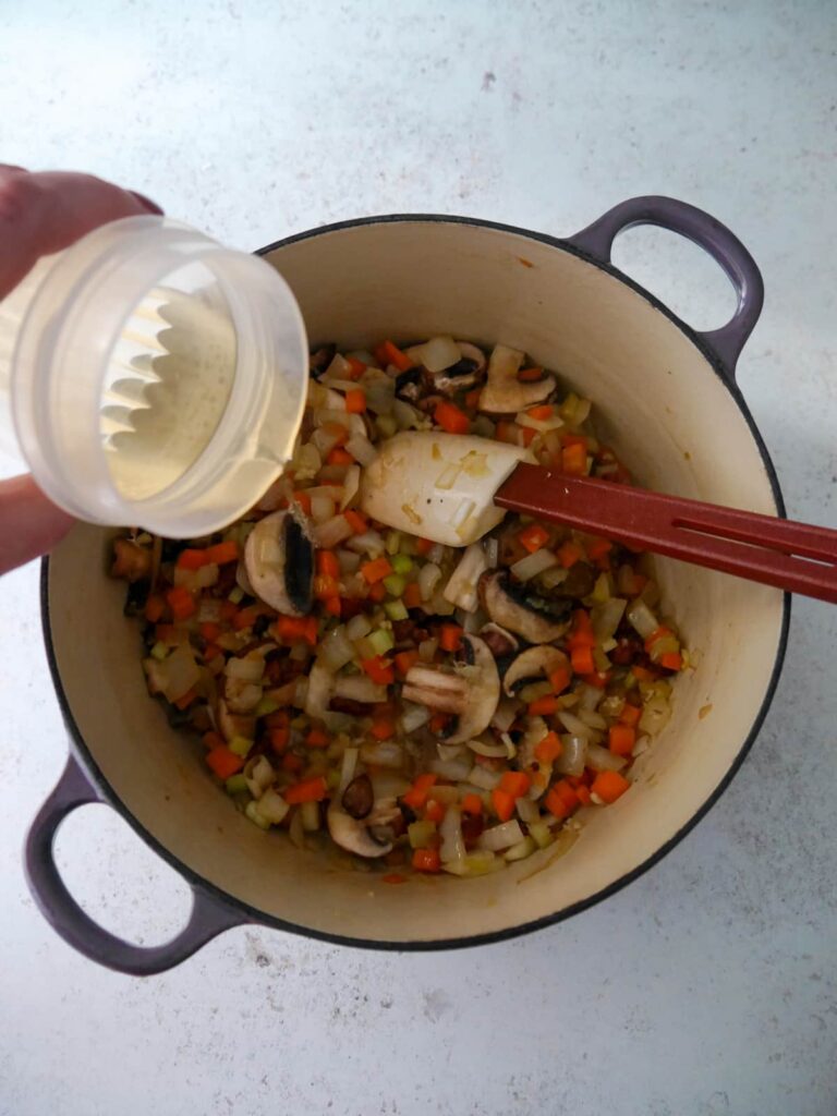 White wine being added to a Dutch oven filled with sauteed onion, carrot, celery and mushroom.