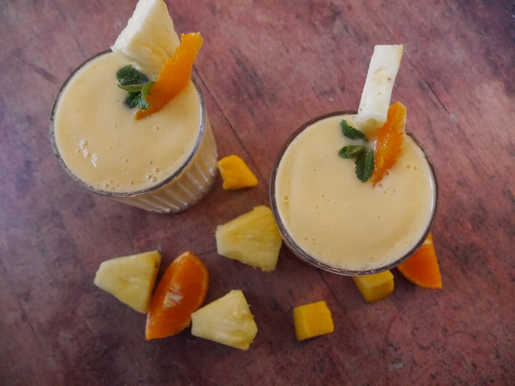 Two glasses of tropical mango pineapple smoothie garnished with a wedge of fresh pineapple and a slice of clementine.